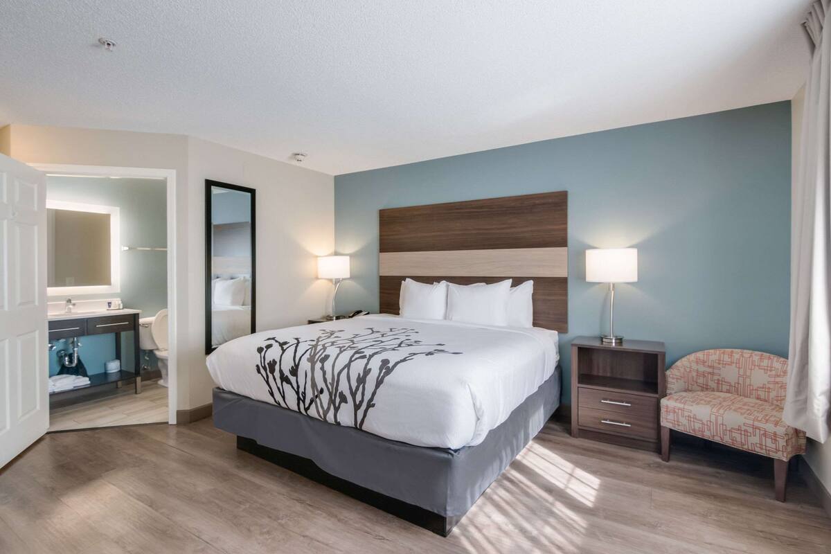 A hotel room with a large bed, bedside tables, a chair, a mirror, and a bathroom visible through an open door.