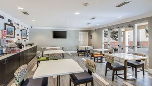 A modern dining area with multiple tables and chairs, a kitchen counter with appliances, and large windows offering views. - Sleep Inn & Suites Columbus near Fort Benning (Columbus)