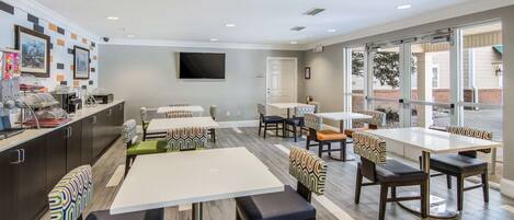 A modern dining area with multiple tables and chairs, a kitchen counter with appliances, and large windows offering views.