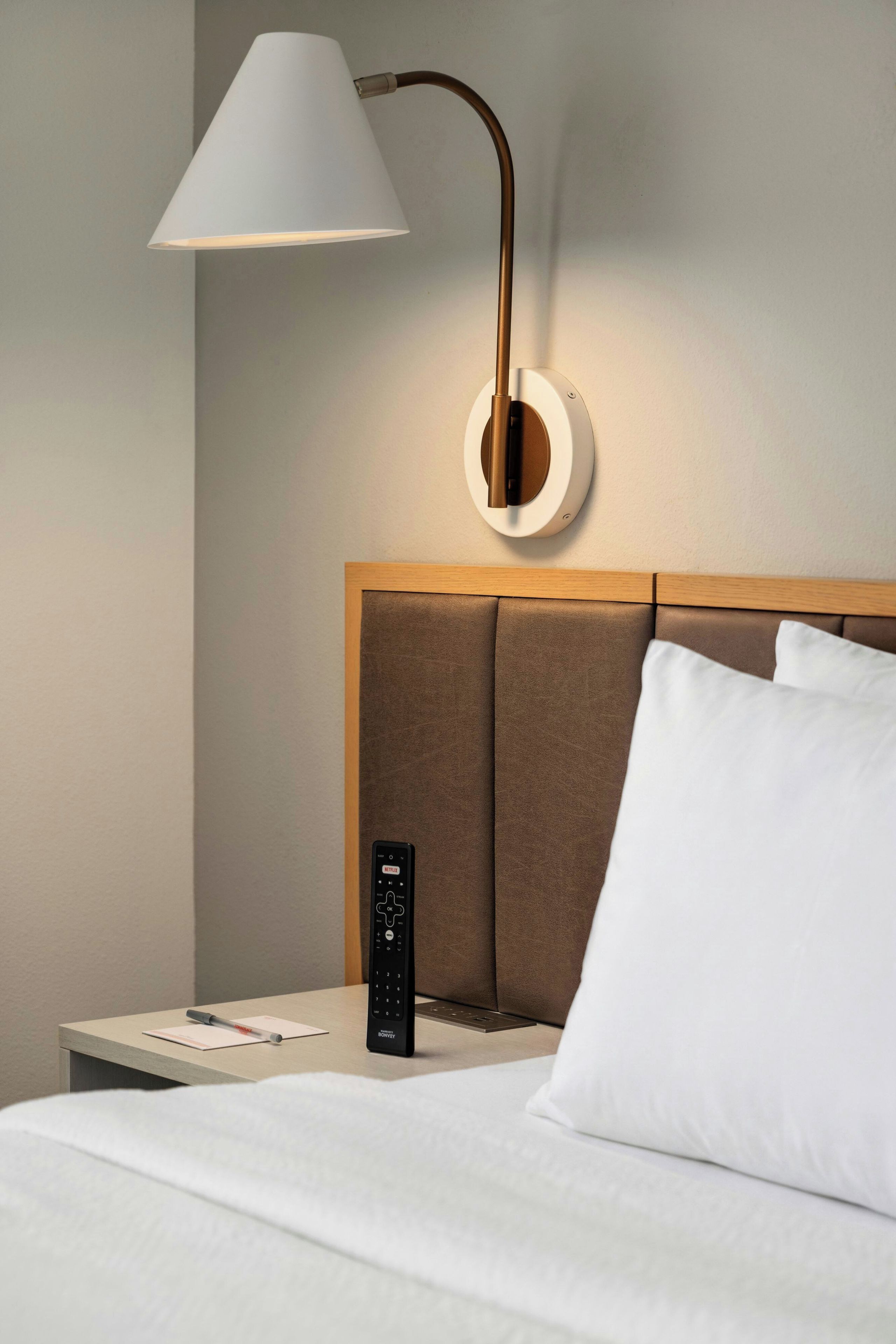 A hotel room with a bed, a nightstand, a remote control, a wall-mounted lamp, and a pillow.