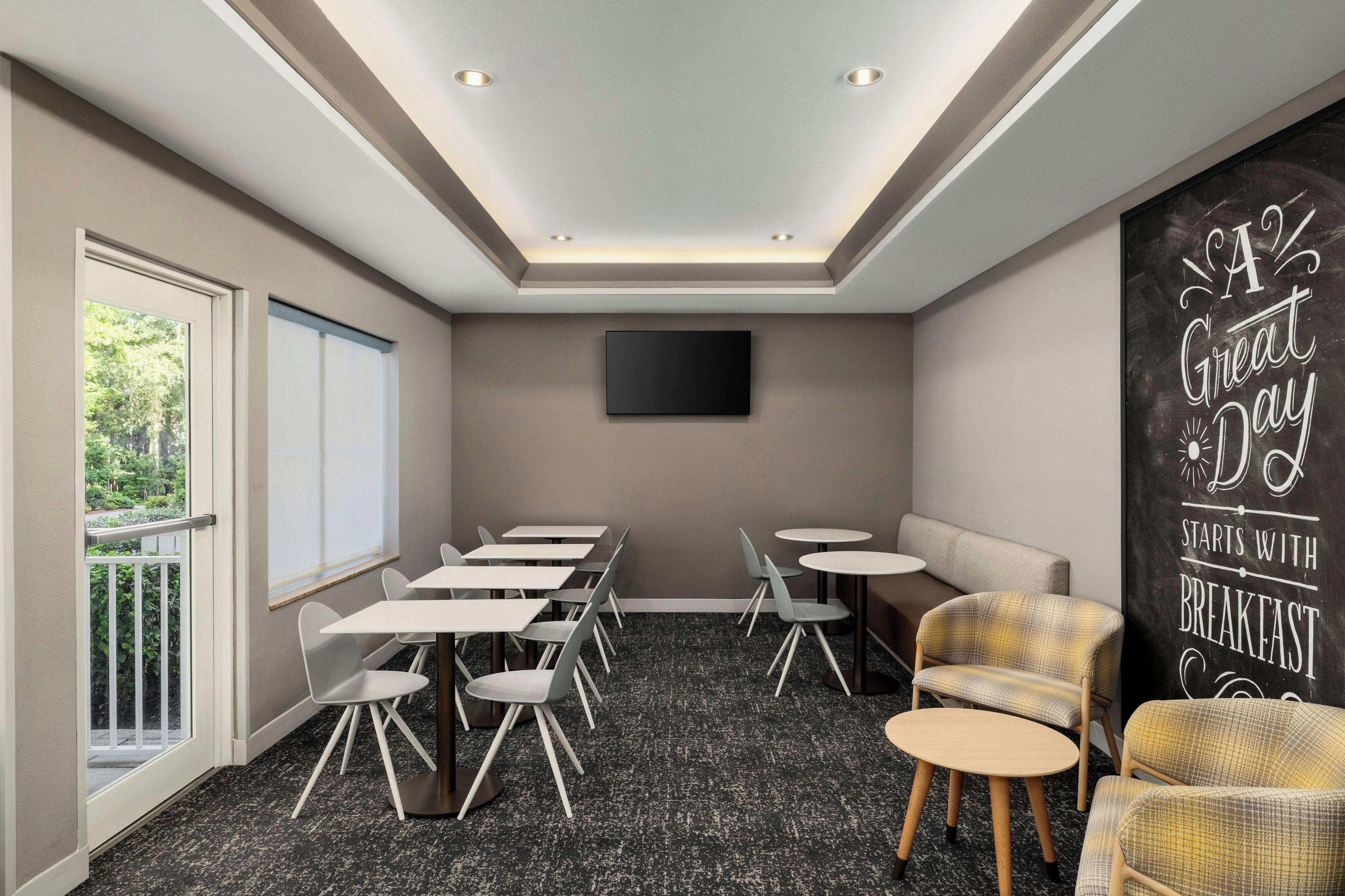A conference room with a flat-screen TV, multiple tables, chairs, and a large window with blinds.