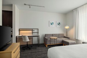 Habitación de hotel con cama, escritorio con silla, sofá, una mesita redonda, televisor y dos cuadros en la pared.