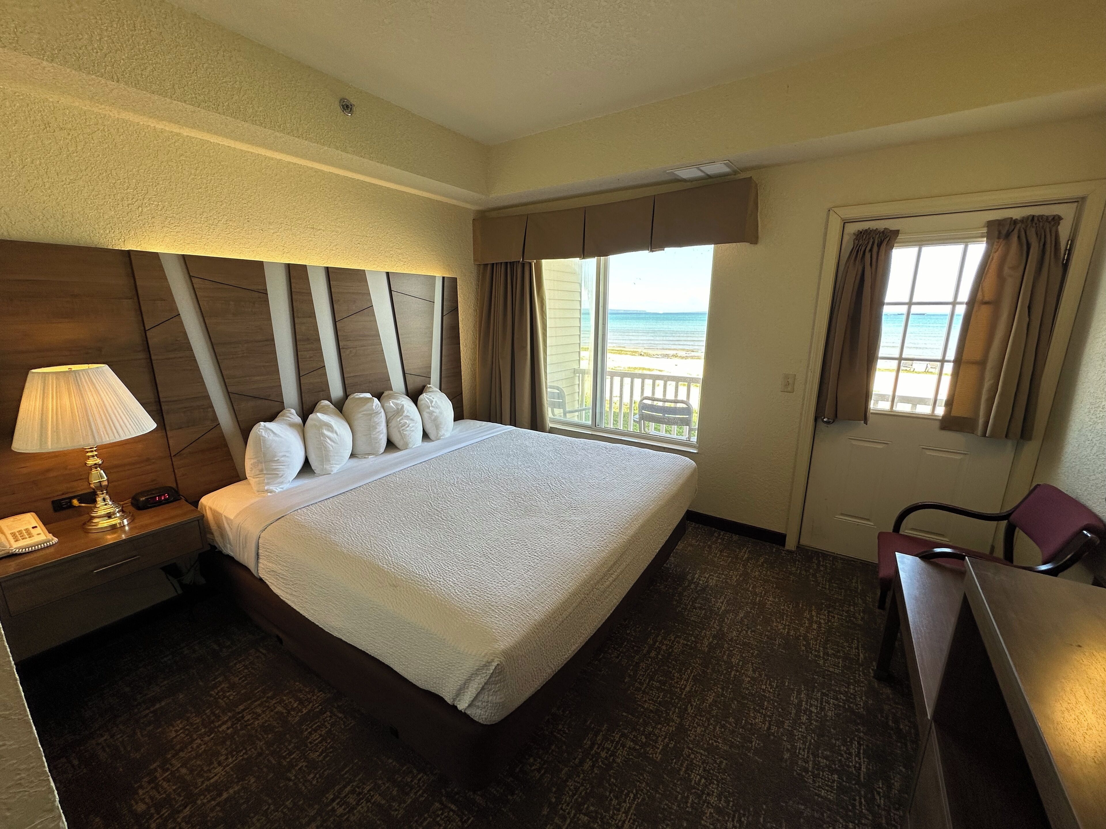 A hotel room with a large bed, a bedside table with a lamp, a chair, and a window with a view of the beach.