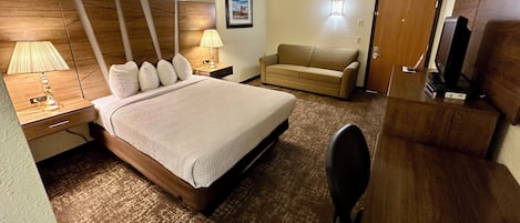 A hotel room with a large bed, a desk, a chair, a sofa, a TV, and a picture on the wall.