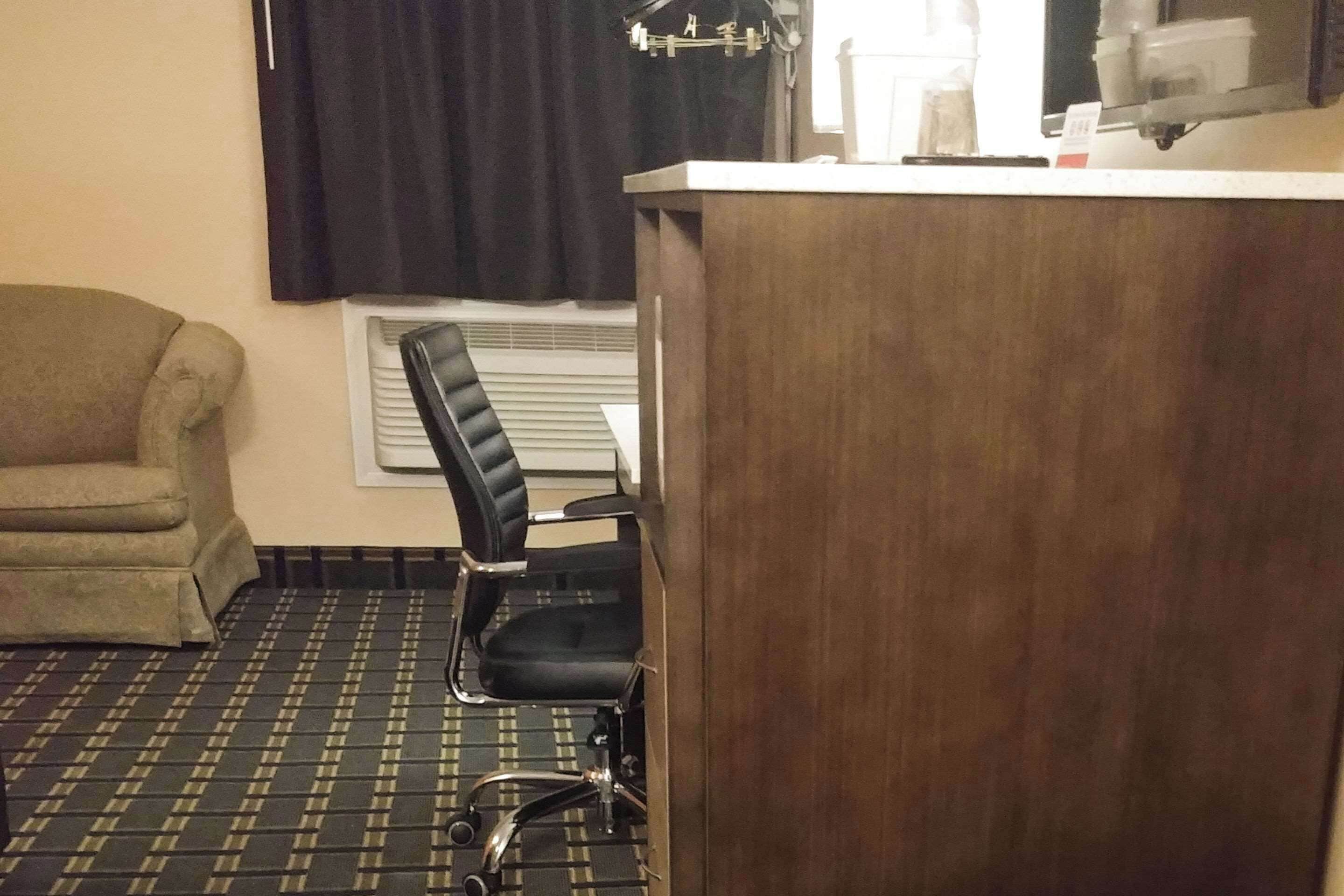 A hotel room with a wooden desk, a black office chair, a beige armchair, and a patterned floor.