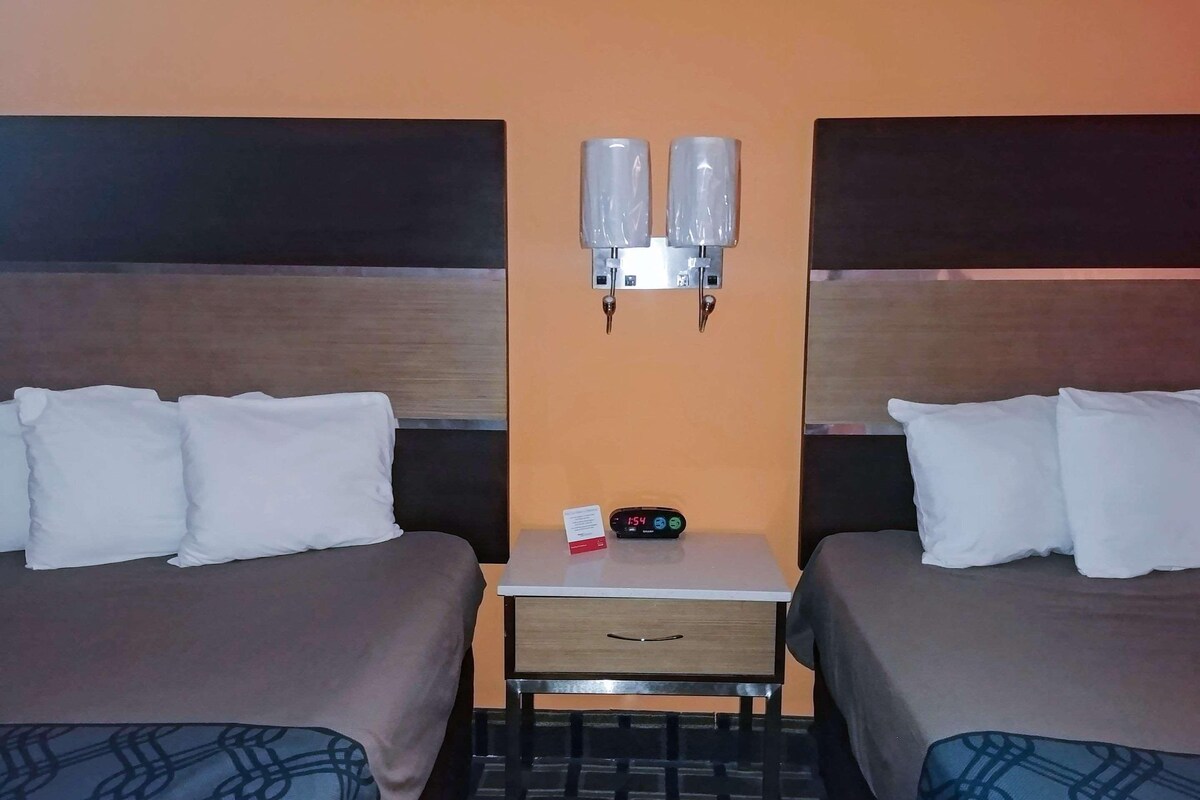 Ein Hotelzimmer mit zwei Betten, einem kleinen Tisch mit einer Uhr und einem Schild sowie einer wandmontierten Lampe.