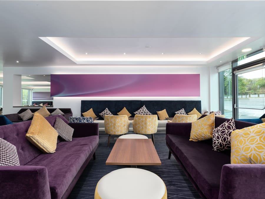A modern lounge area with purple sofas, yellow patterned cushions, and a large wall-mounted screen.