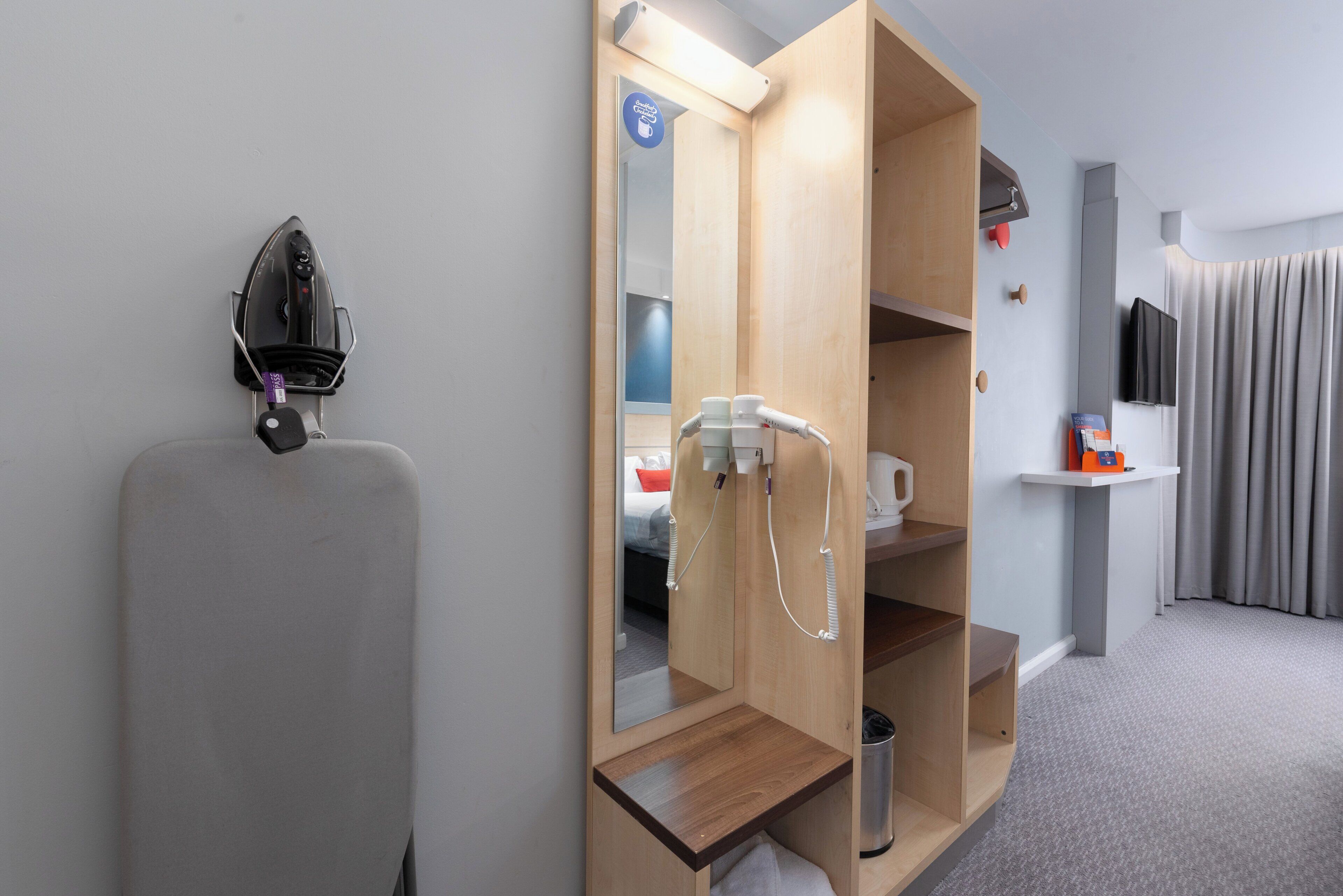 Ein Hotelzimmer mit integriertem Fön, einem Spiegel, einer Ablagefläche mit Kaffeemaschine und einem an der Wand montierten Fernseher.