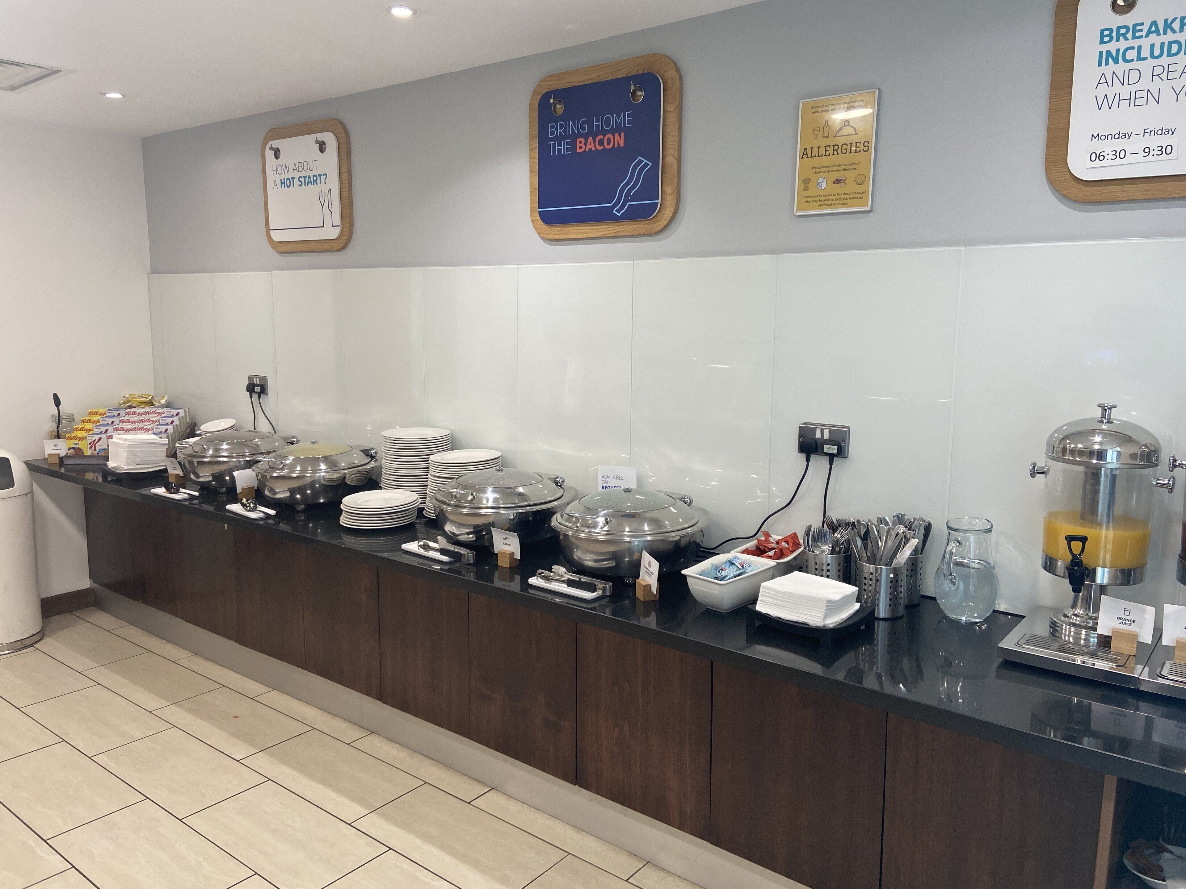 A buffet setup with various food containers, utensils, and a sign indicating breakfast hours.
