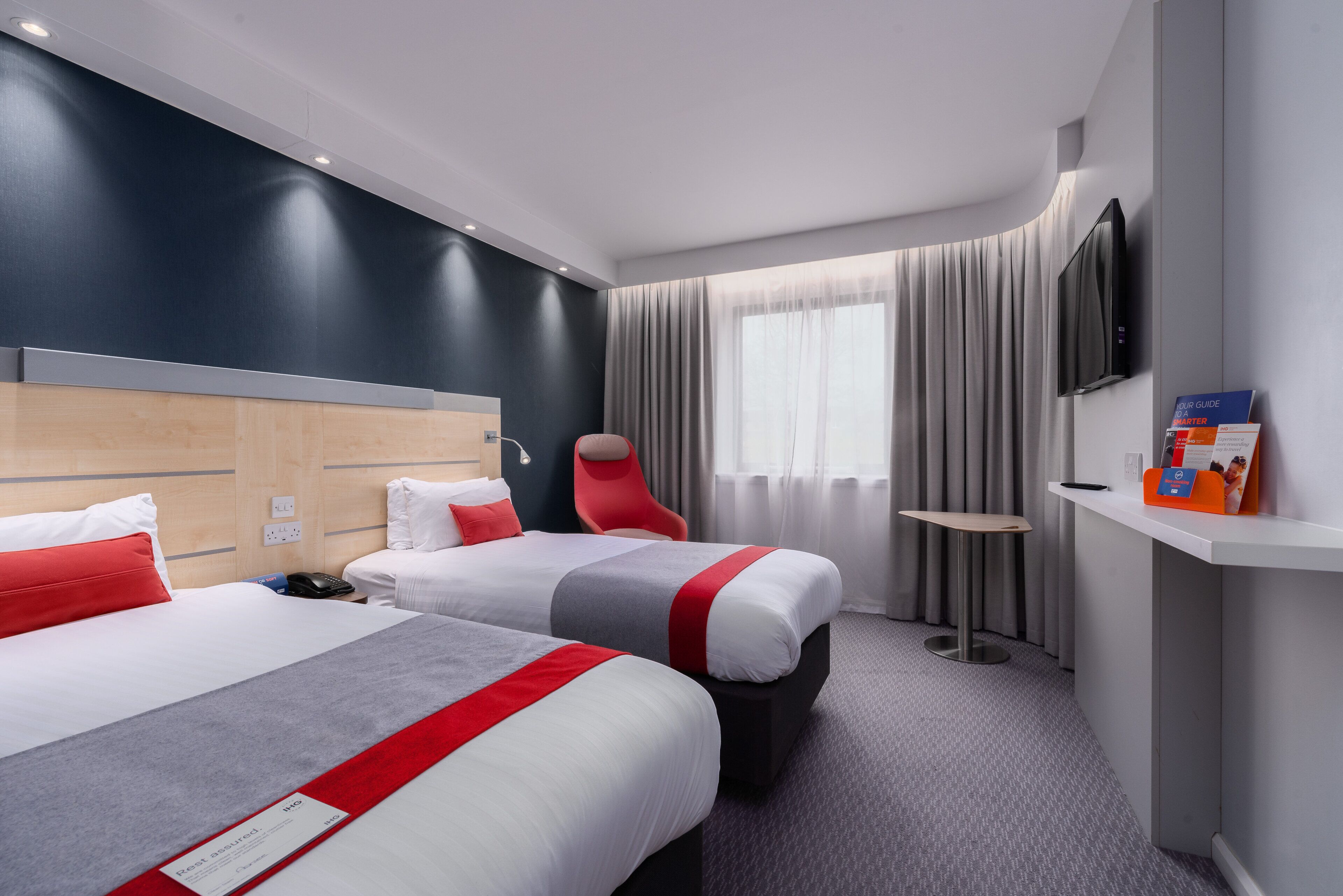 Habitación de hotel con dos camas, una silla roja, una mesita, televisión y mostrador de recepción.