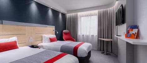 Habitación de hotel con dos camas, una silla roja, una mesita, televisión y mostrador de recepción.