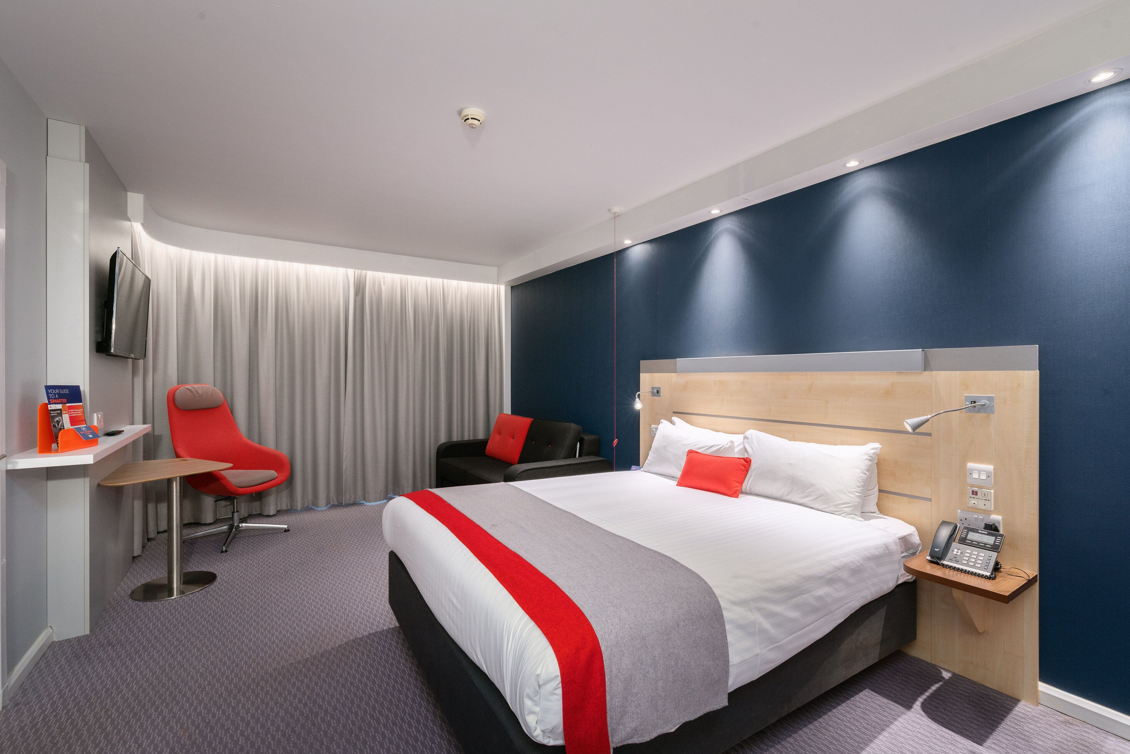 A modern hotel room with a large bed, a desk with a chair, a TV, and a telephone.