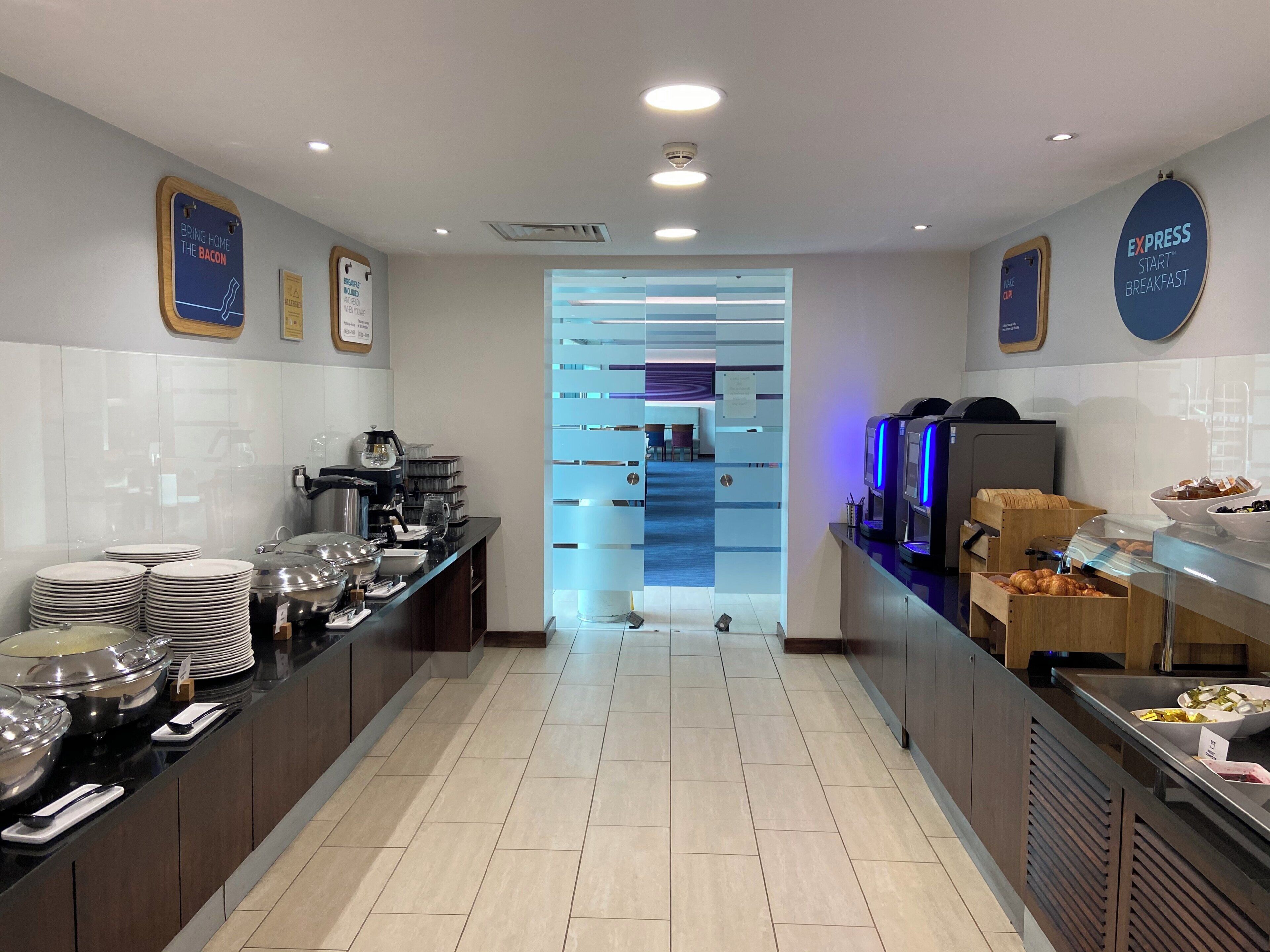 A modern hotel breakfast area with buffet counters, self-service beverage dispensers, and a clear view of the dining area.