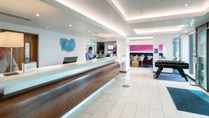 Ein Hotel-Rezeptionsbereich mit einem Holzbüro, einem digitalen Bildschirm und einem blauen Schild. Im Hintergrund befinden sich eine Couch und ein Tisch.