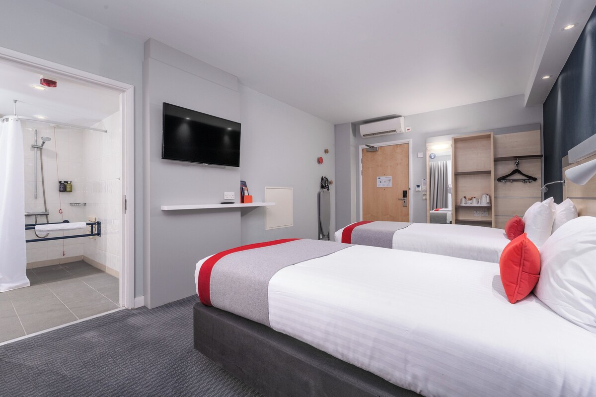 A hotel room with two beds, a TV, a shower, and a small bathroom.