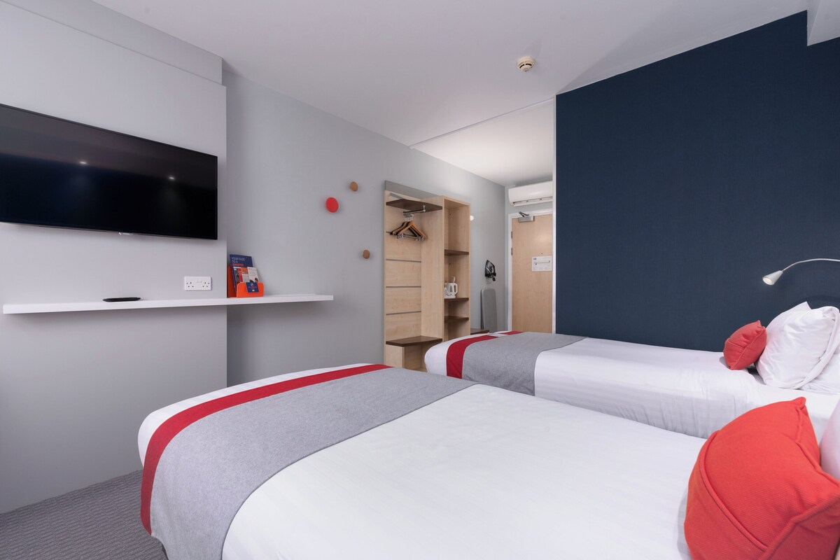 A hotel room with two beds, a TV mounted on the wall, a wardrobe, and a small shelf with items.