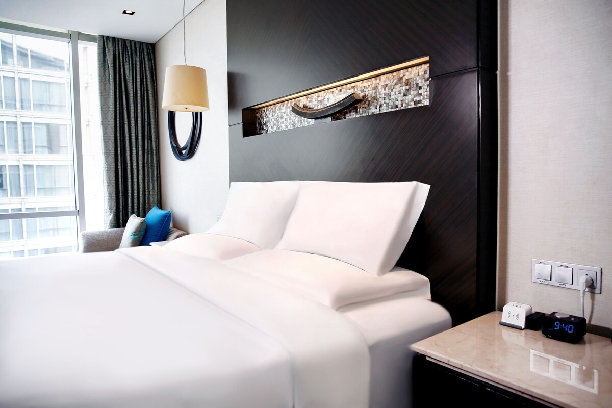 A hotel room with a large bed, a bedside table with a clock, and a window with curtains.