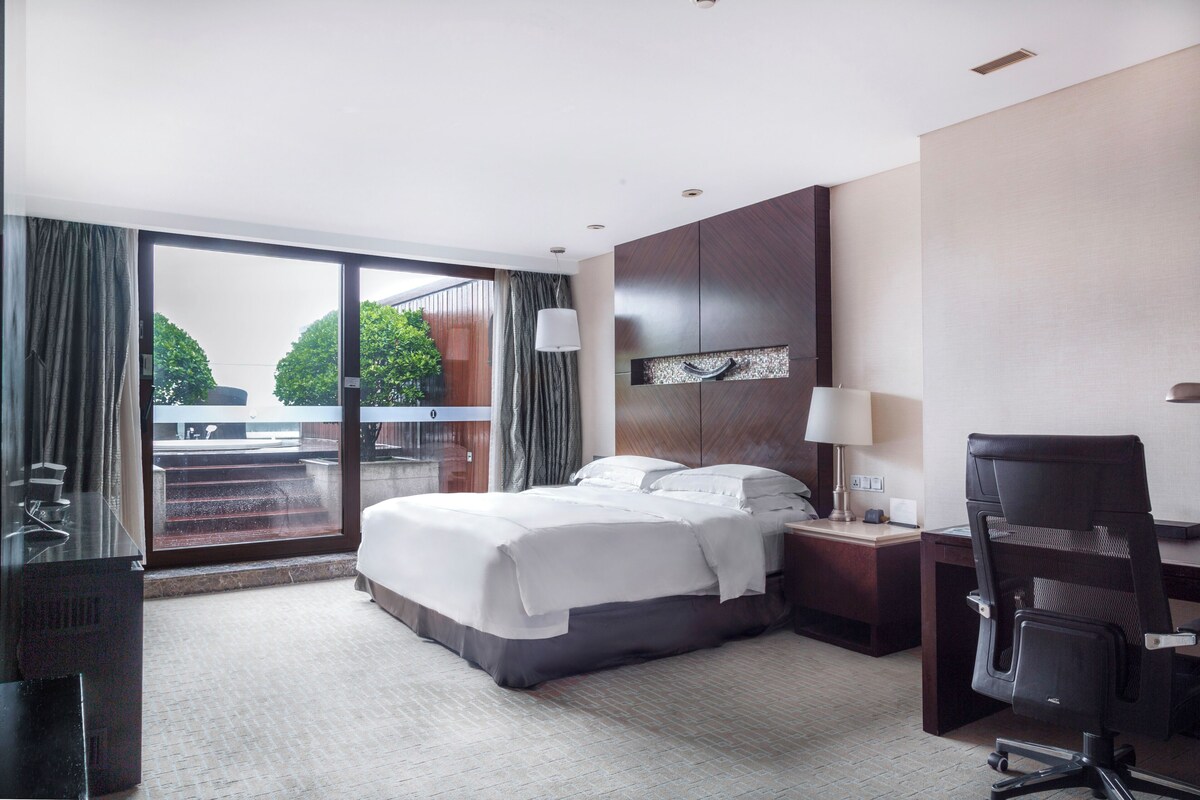 A hotel room with a large bed, a desk, a chair, a desk lamp, and a view of the outdoors through a sliding door.