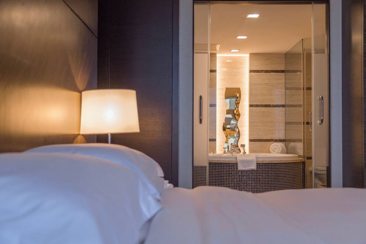 A hotel room with a bed, a lamp, a bathroom with a shower, and a mirror.