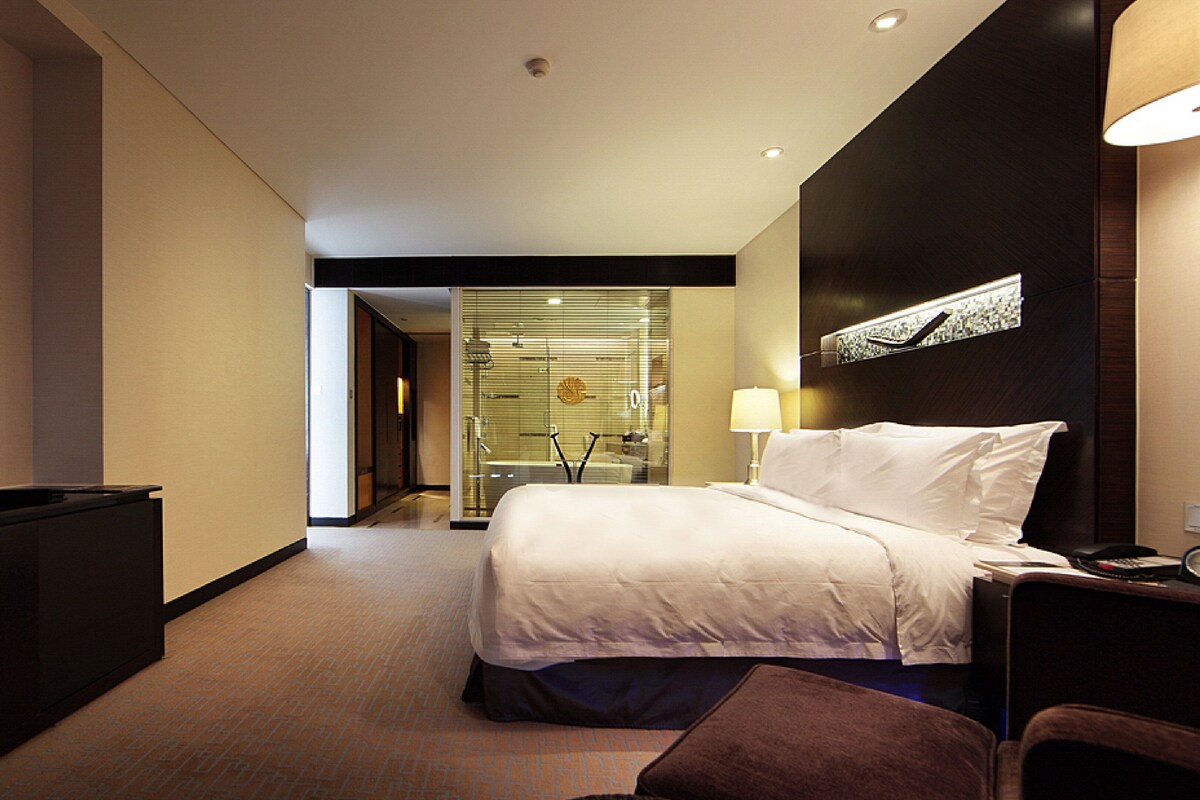 A hotel room with a bed, a TV, a desk, and a bathroom visible through a glass door.
