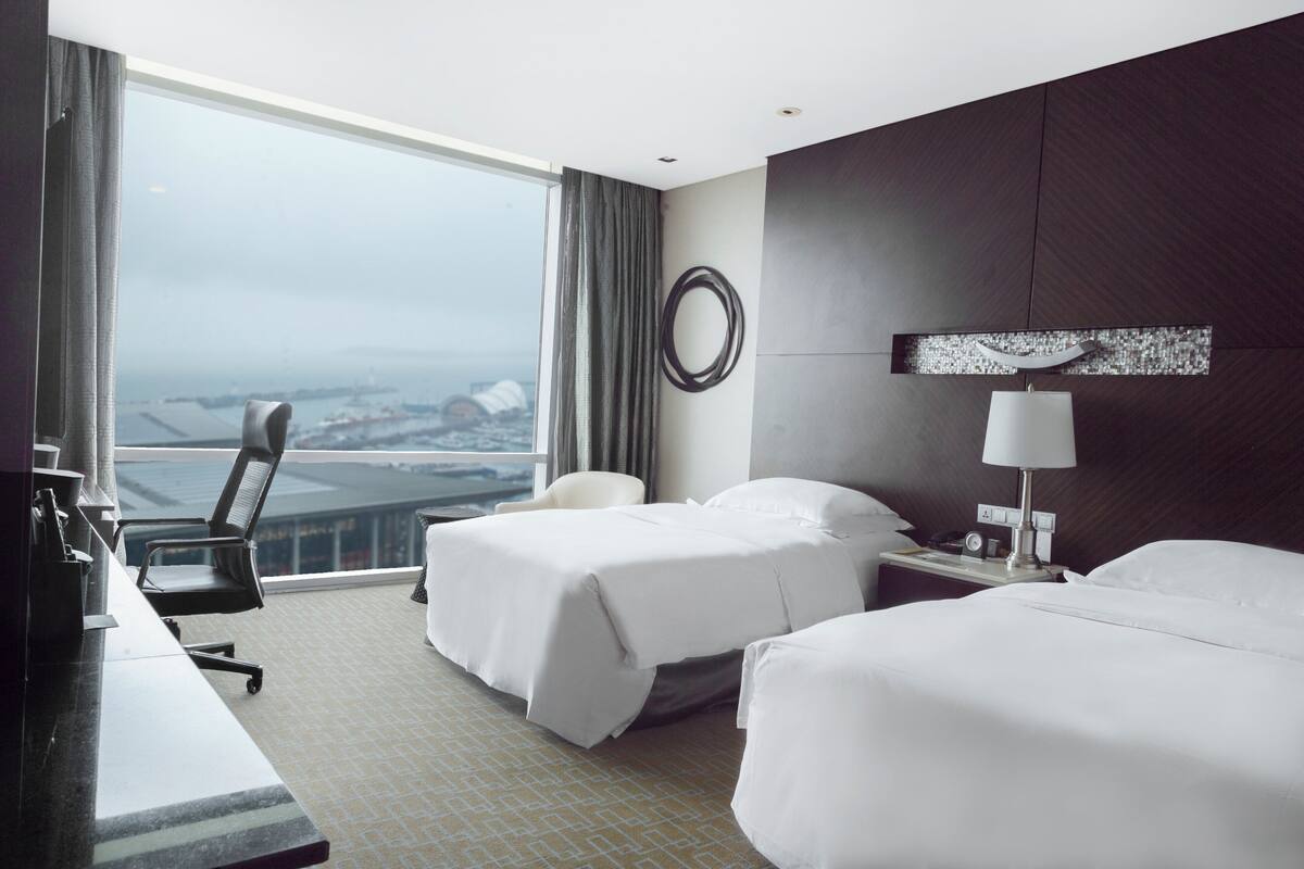 A hotel room with two beds, a desk, a chair, a large window with a view, and a wall-mounted lamp.