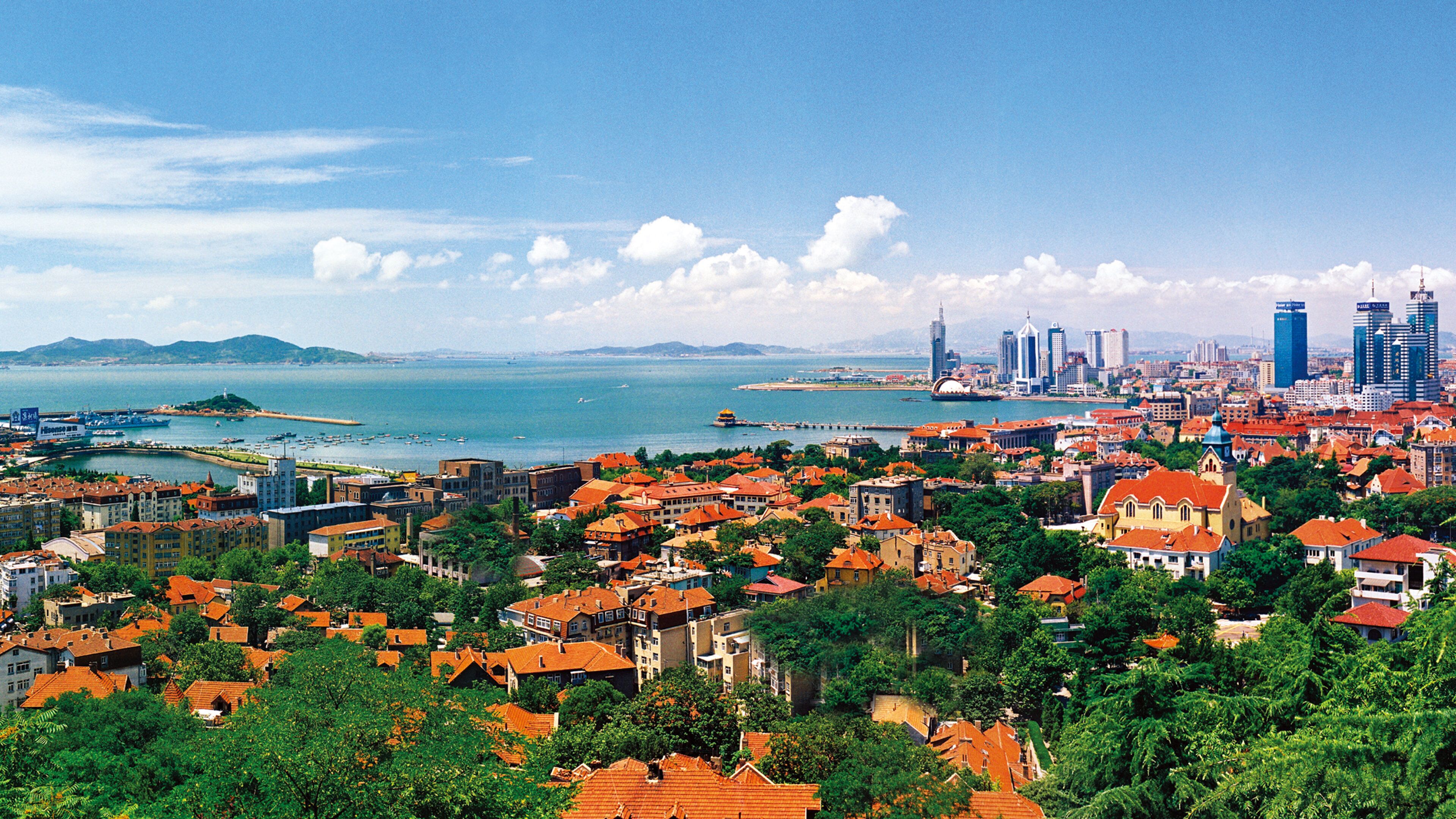 A coastal cityscape with dense urban buildings, a harbor with boats, and a clear blue sky.