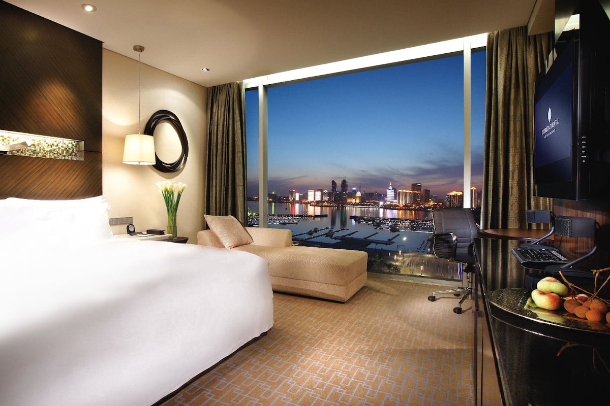 A hotel room with a large bed, a sofa, a desk with a computer, and a view of a cityscape at dusk.
