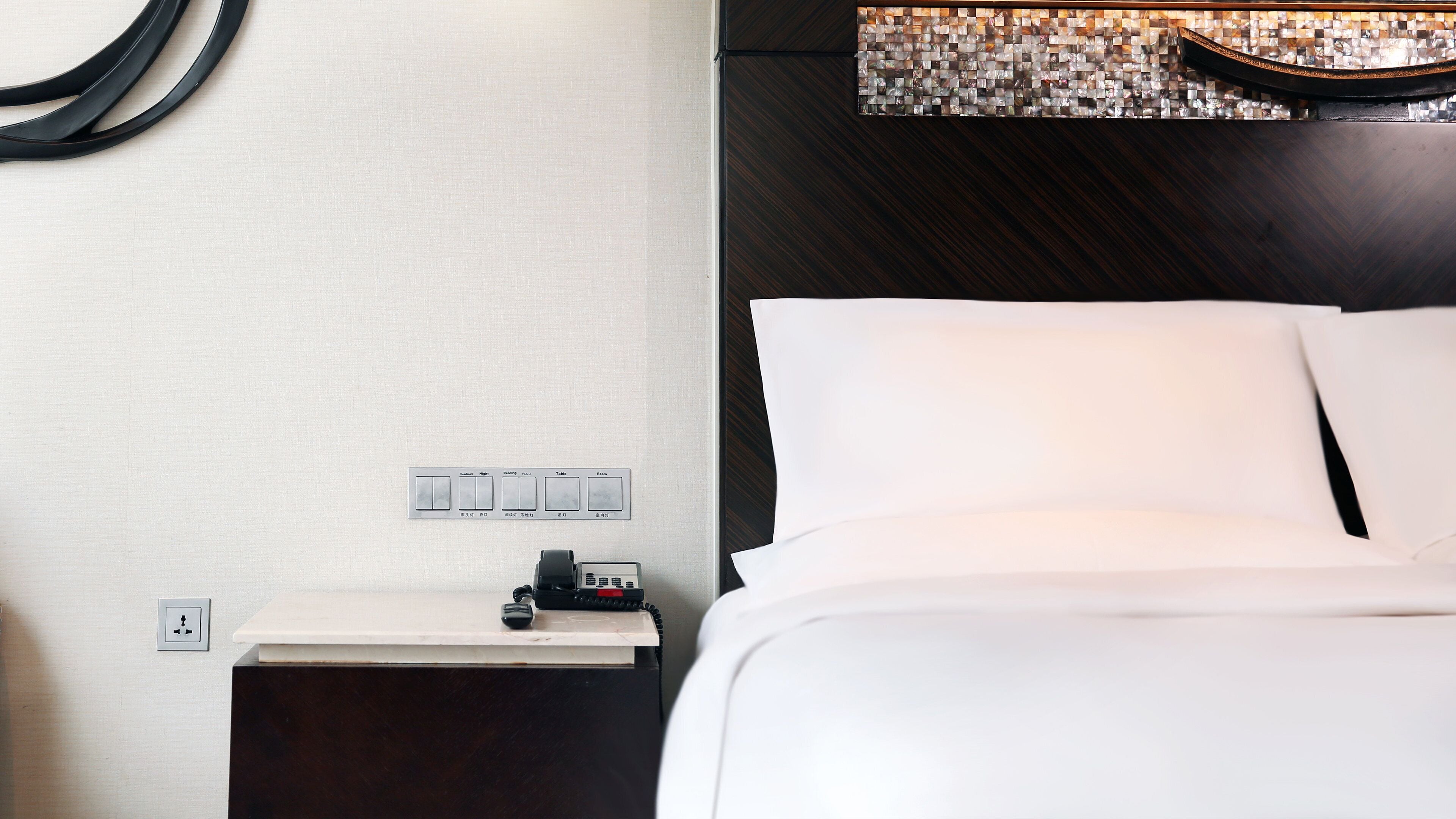 A hotel room with a bed, a nightstand with a phone, and a wall-mounted light switch panel.