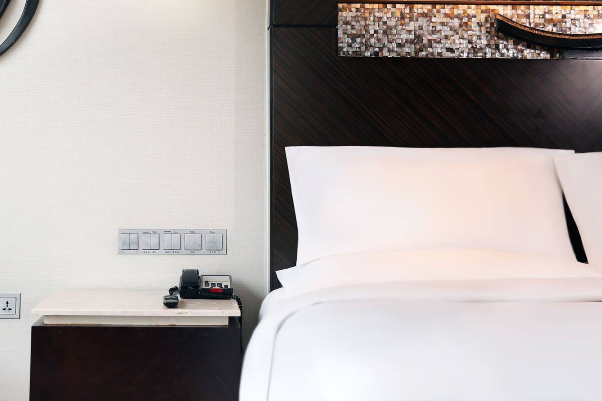 A hotel room with a bed, a nightstand with a phone, and a wall-mounted light switch panel.