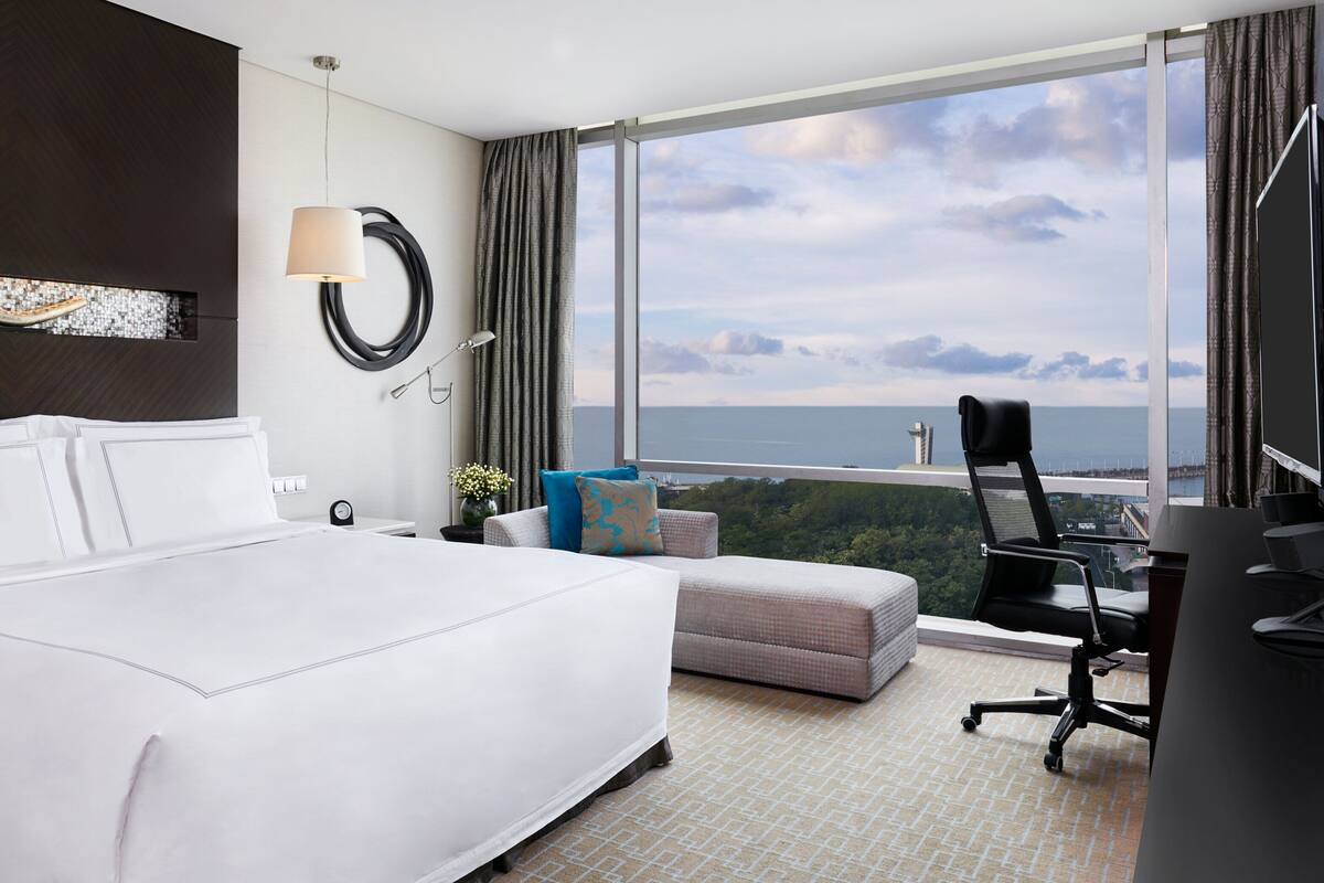 A hotel room with a large bed, a desk, a chair, a lamp, a view of the ocean, and a window with curtains.