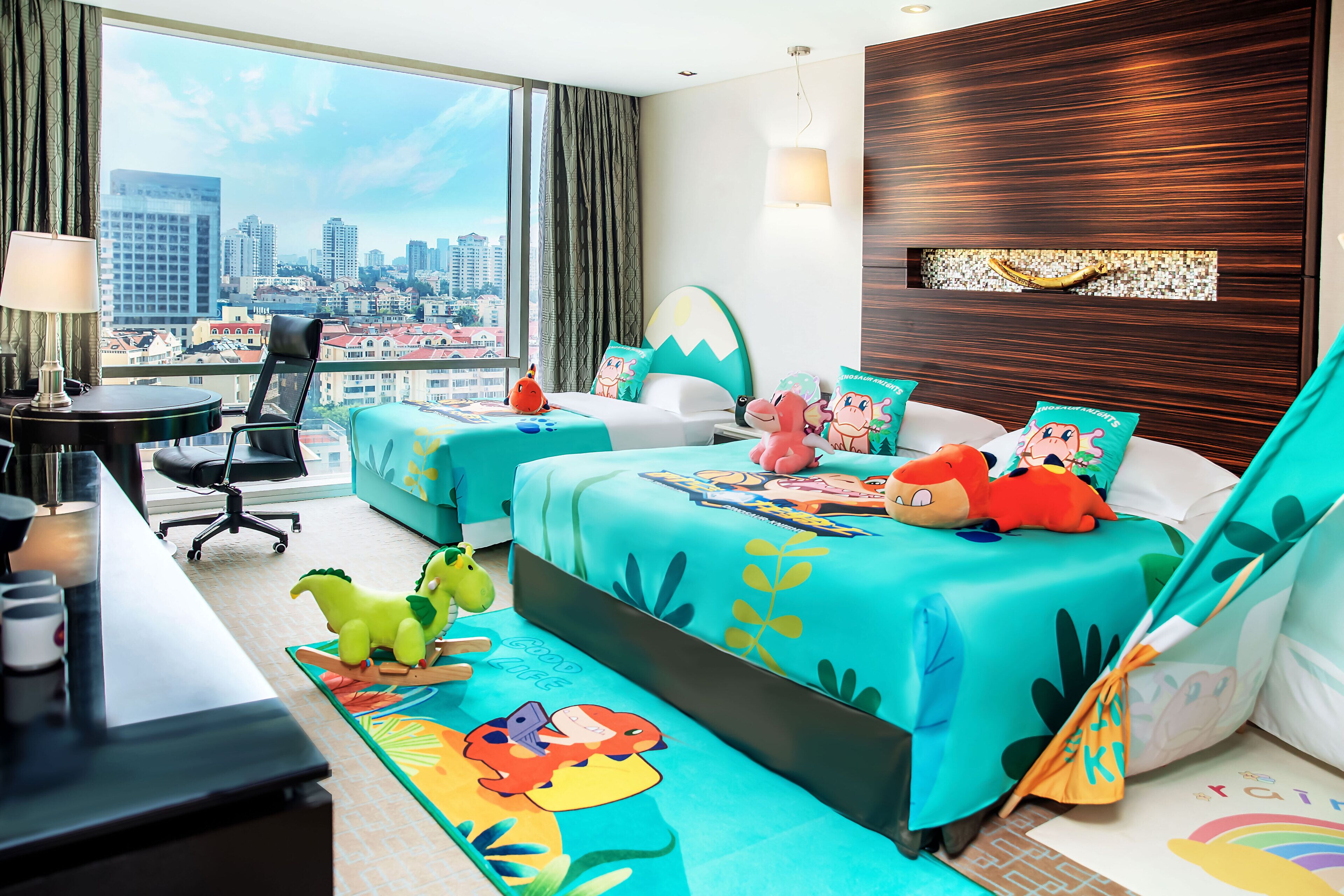 A hotel room with a city view, a desk with a lamp, two beds with colorful bedding, and a dinosaur-themed rug.