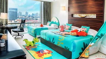 A hotel room with a city view, a desk with a lamp, two beds with colorful bedding, and a dinosaur-themed rug.