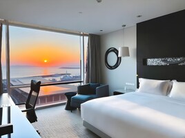 A hotel room with a large bed, a desk, a chair, and a view of the sunset over the water.