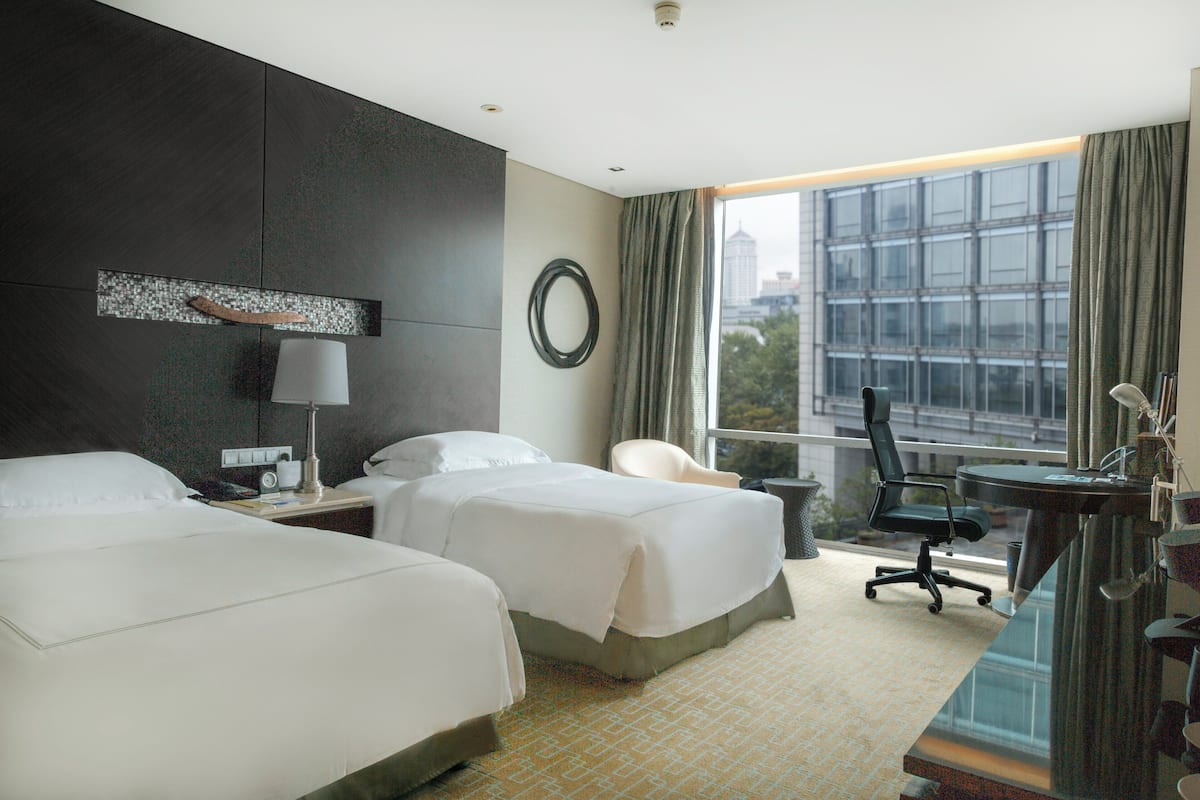 A hotel room with two beds, a desk, a chair, a lamp, and a view of the city through large windows.