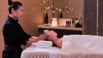 A person receiving a massage in a room with a bed, a massage table, and a wall-mounted shelf with various items.