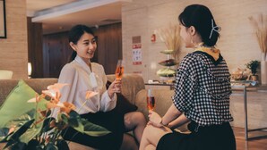 Two people sitting on a couch in a lobby with a plant and a small table with food.