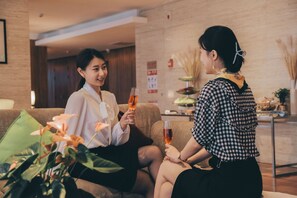 Zwei Personen sitzen auf einer Couch in einer Lobby, mit einer Pflanze und einem kleinen Tisch mit Essen.