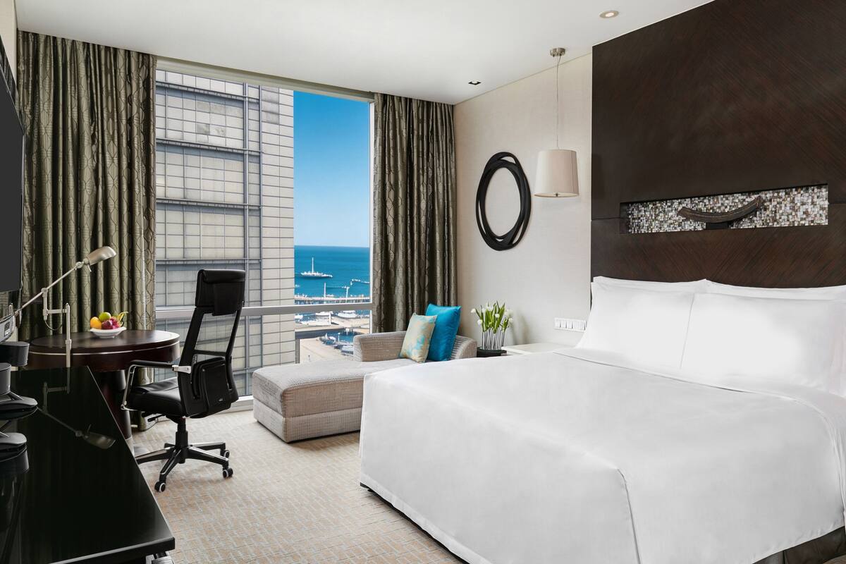 A hotel room with a large bed, a desk with a chair, a small sofa, a view of the sea, and a cityscape through the window.