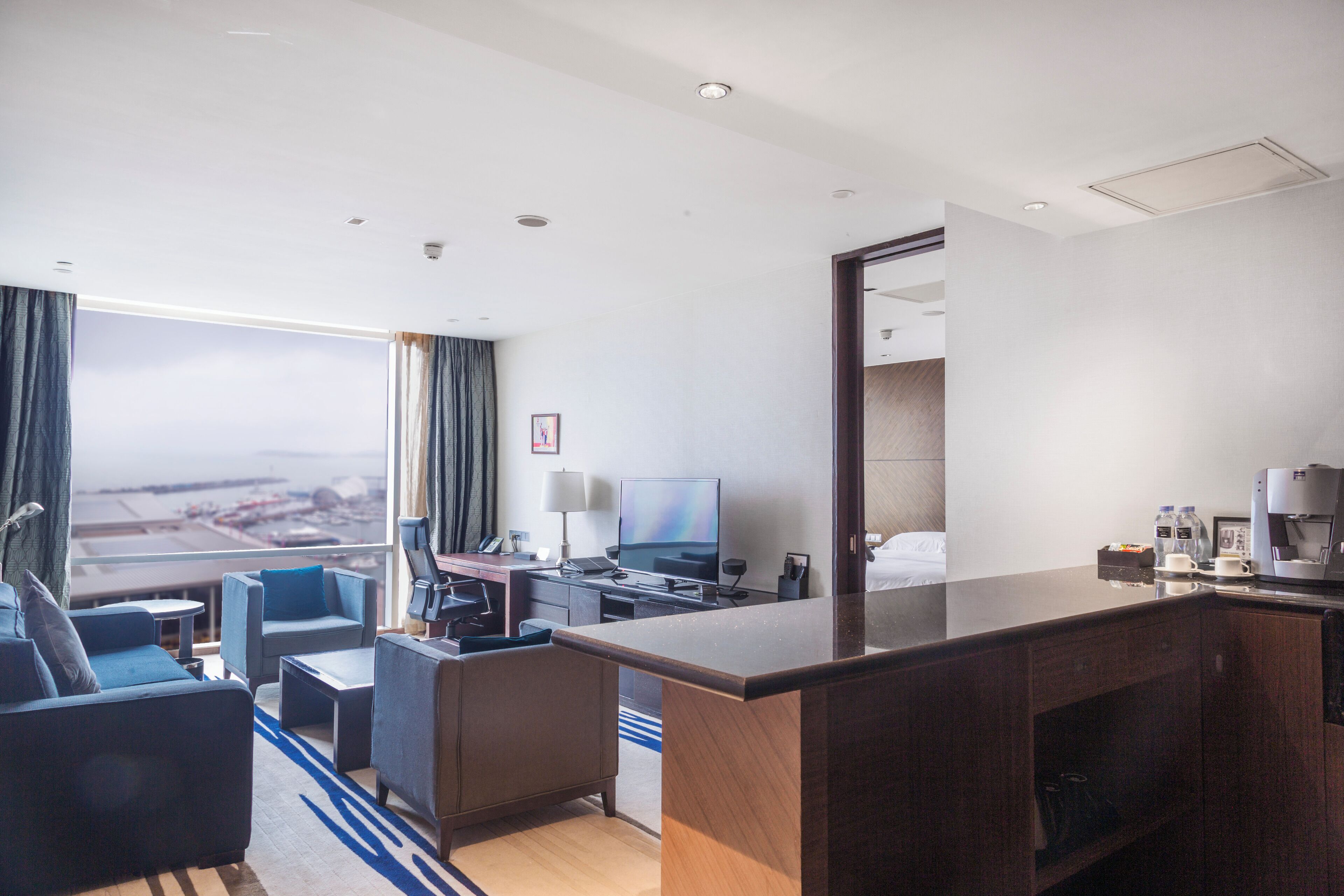 A hotel room with a large window, a desk, a television, and a coffee machine.