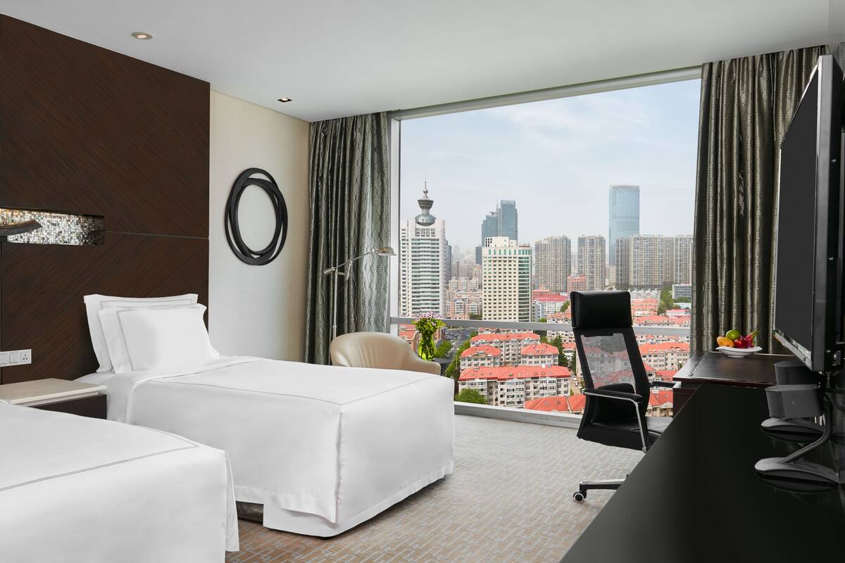 A hotel room with two beds, a desk, a chair, a large window with a city view, and a TV.