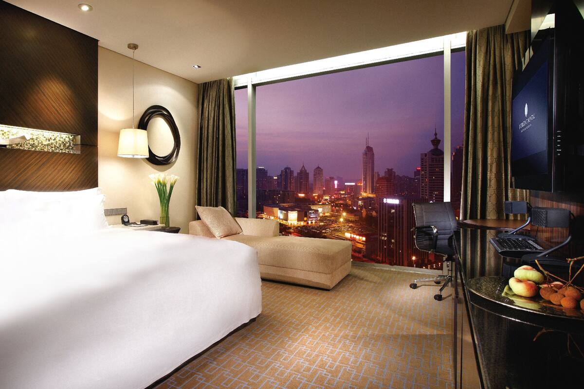 A hotel room with a large bed, a sofa, a desk with a computer, and a view of a cityscape at dusk.