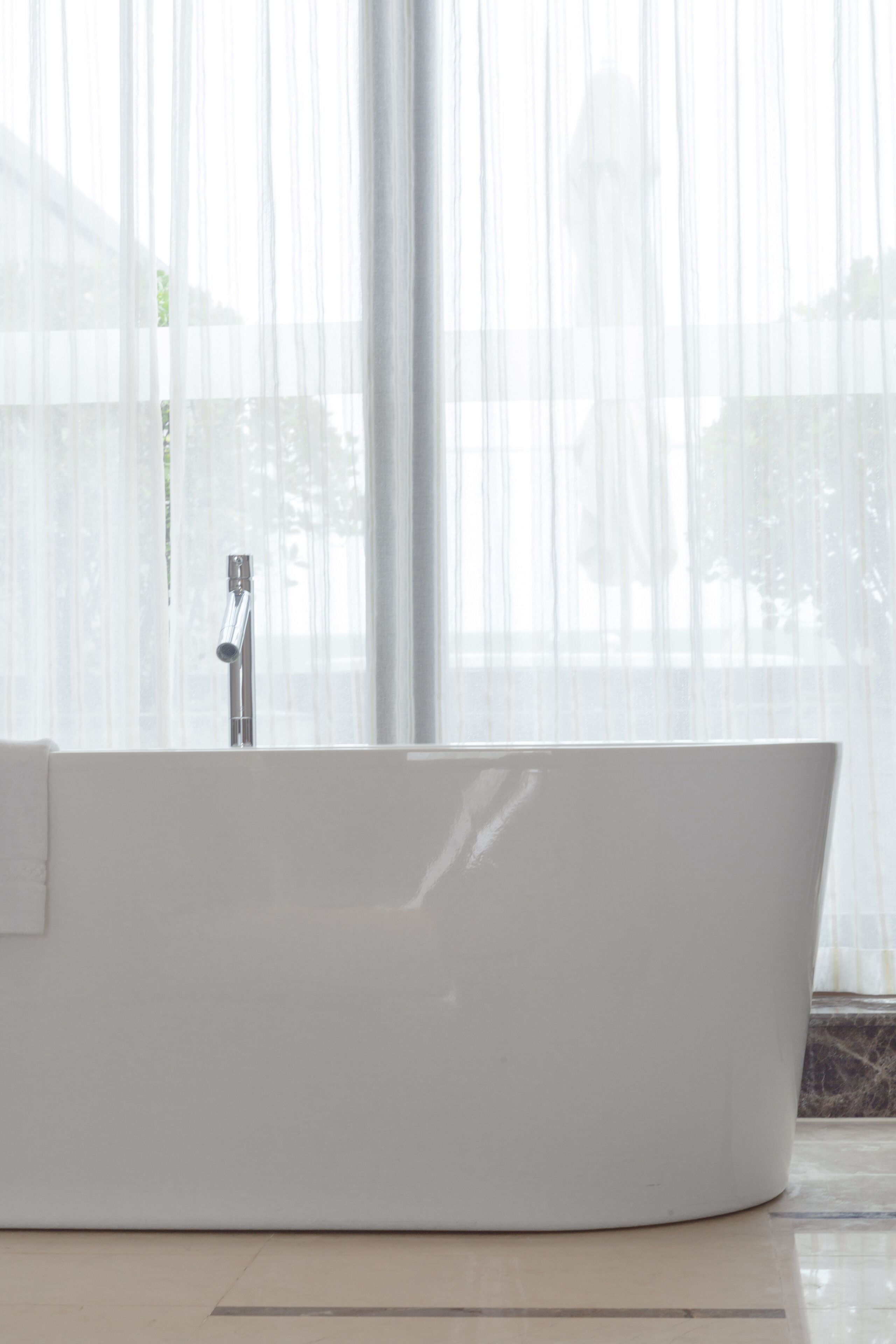 Un baño moderno con una bañera independiente, un grifo a la vista y un amplio ventanal con cortinas de gasa.
