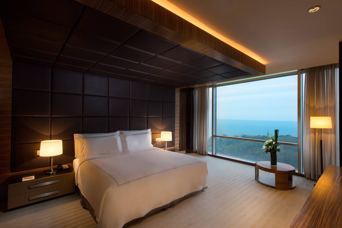 A modern hotel room with a large bed, two bedside lamps, a nightstand, a round wooden table with a vase, and a large window offering a view of the ocean.