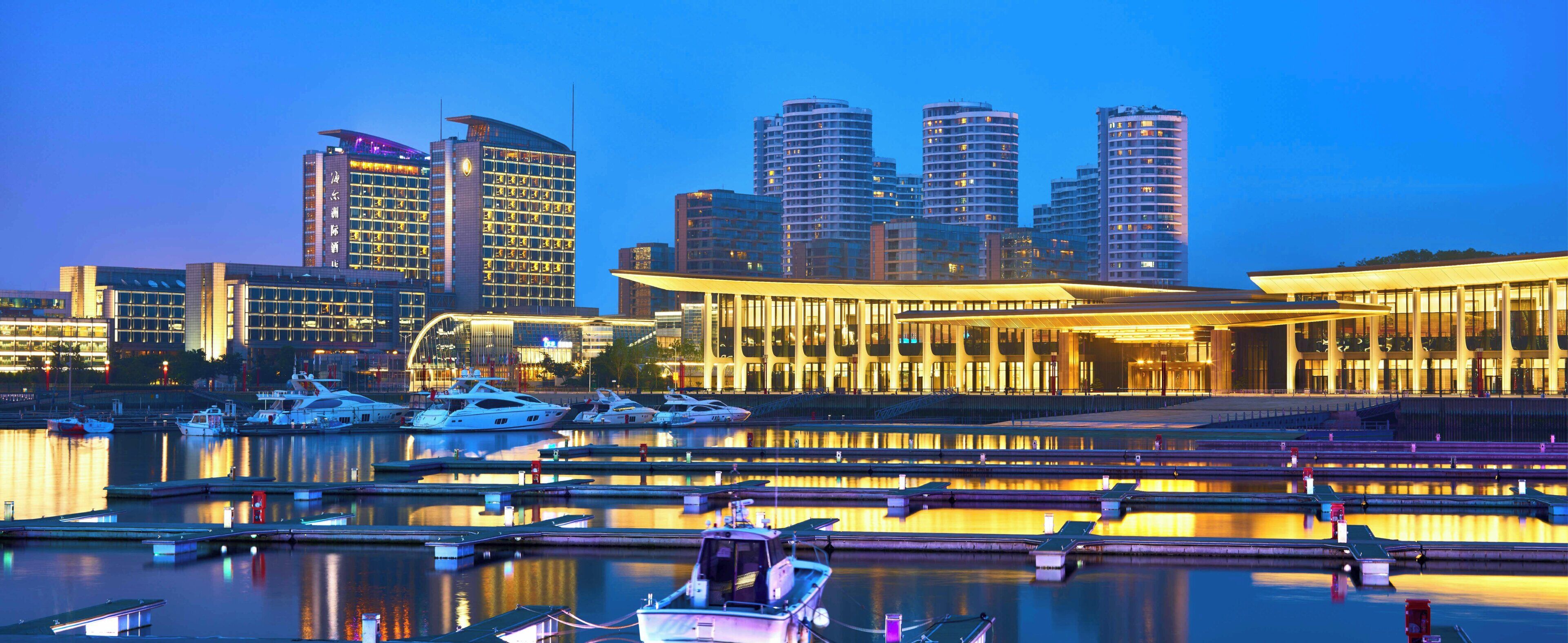 A marina with boats docked, a modern building with large windows, and high-rise buildings in the background.