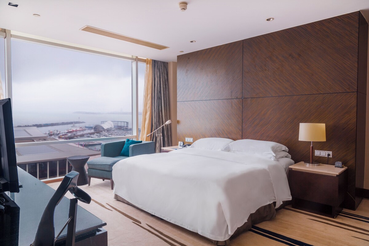 A hotel room with a large bed, a desk, a chair, a TV, and a view of a cityscape.