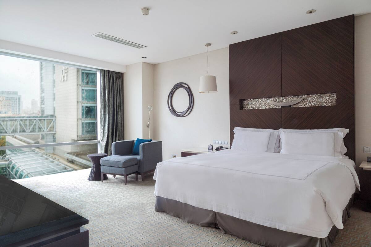 A modern hotel room with a large bed, a seating area with a chair, a desk, and a view of a cityscape through large windows.