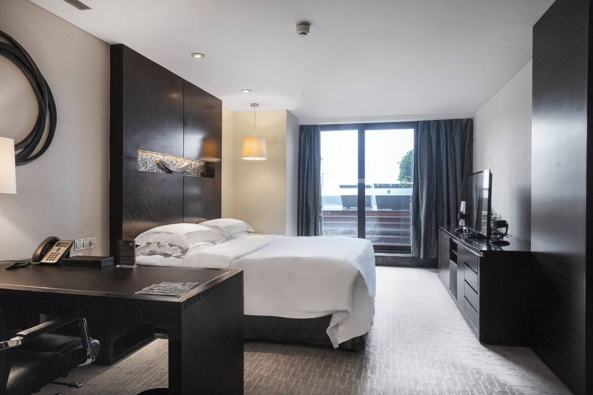 A hotel room with a bed, desk, chair, TV, and a large window with curtains.