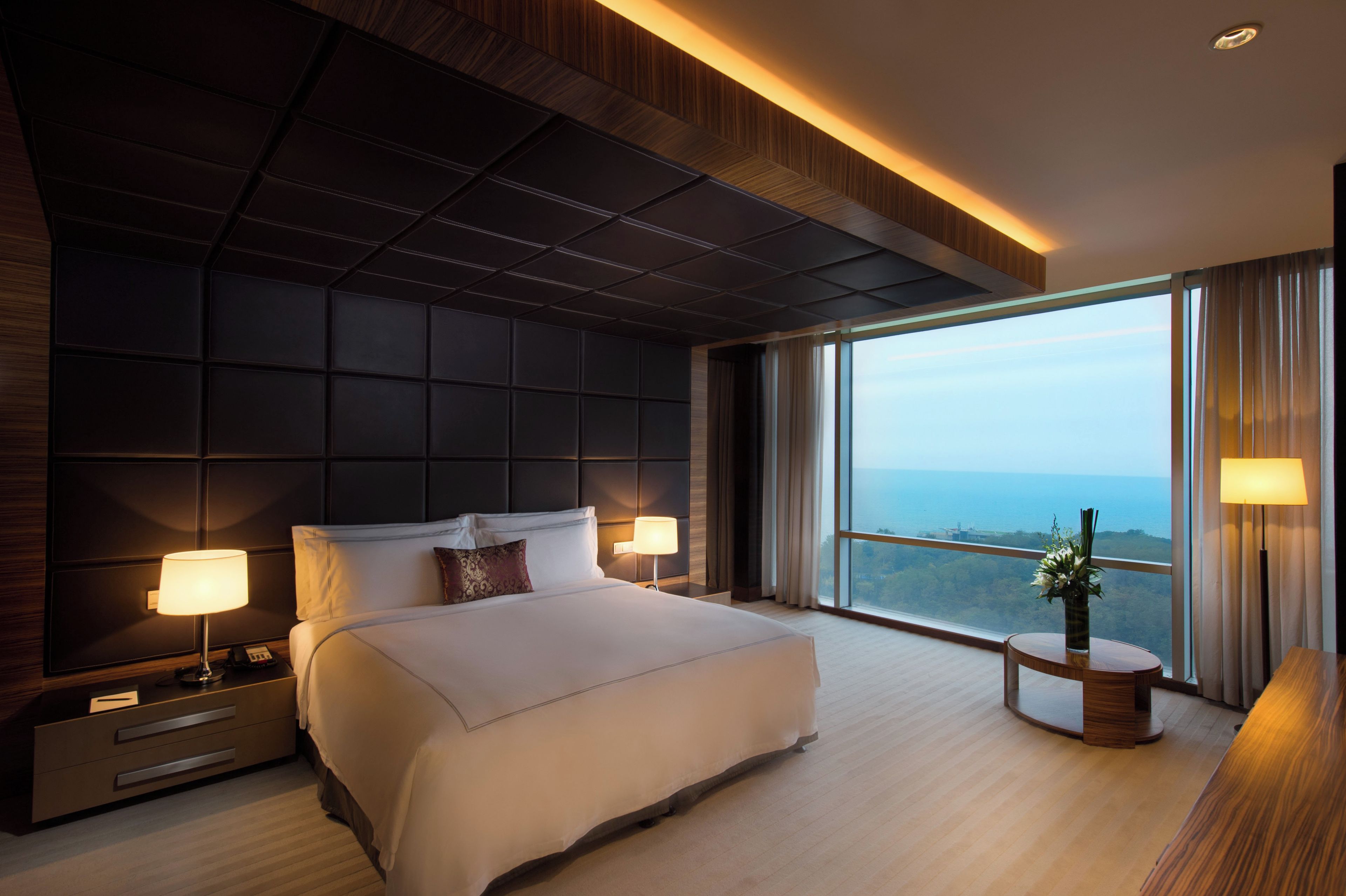Une chambre d’hôtel moderne avec un grand lit, deux lampes de chevet, une table de chevet, une table ronde ornée d’un vase, et une vue sur la mer.