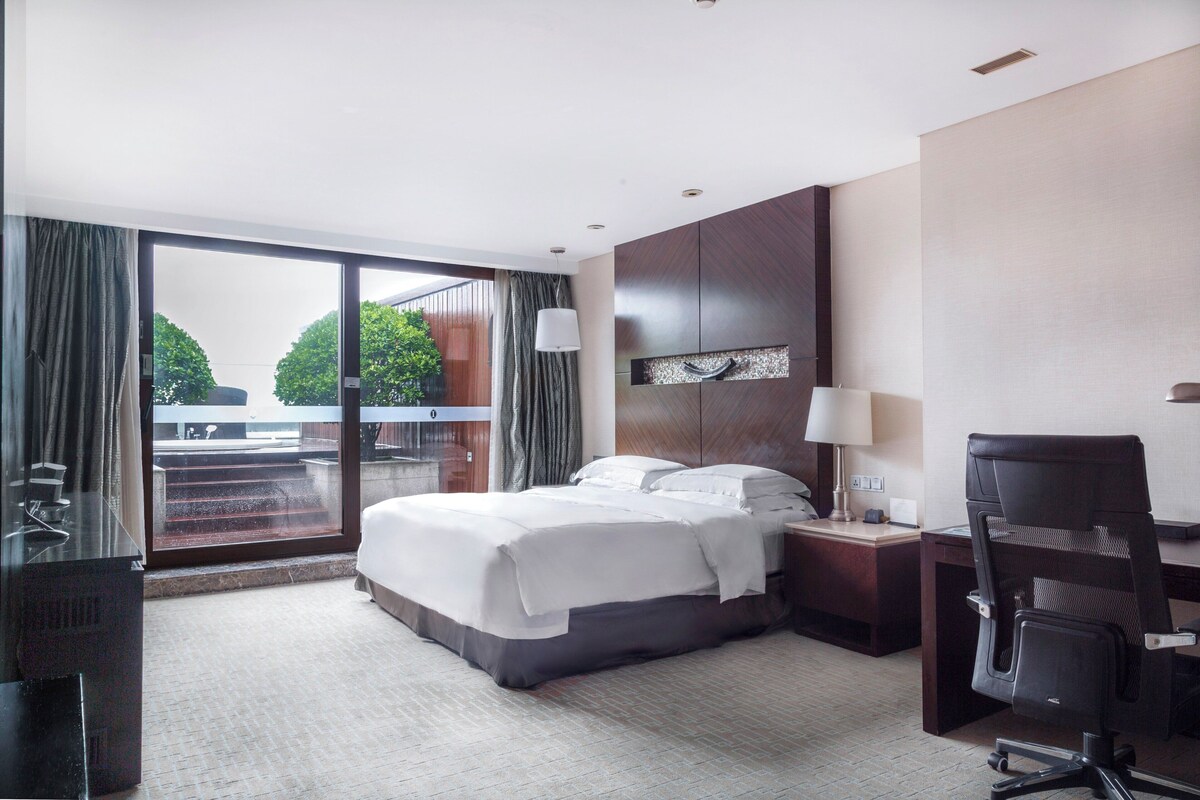 Ein Hotelzimmer mit einem großen Bett, einem Schreibtisch, einem Stuhl und Blick ins Freie.