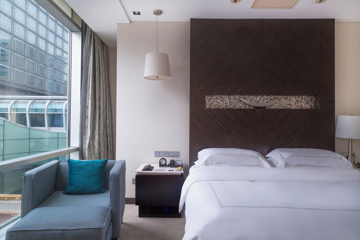 A hotel room with a bed, a desk with a lamp, a chair, a window with a view of a building, and a wall with a decorative panel.